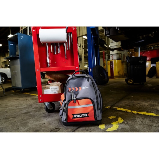A PROTO branded backpack with orange and black colors is placed on the floor next to a red tool cart in an industrial workshop setting.