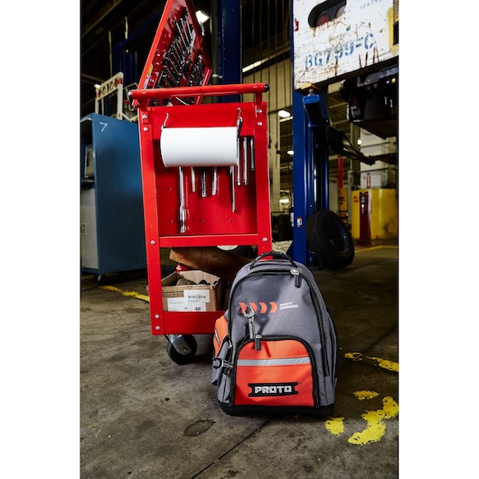 PROTO branded backpack placed in front of a red tool cart equipped with various hand tools and a roll of paper towels in an industrial workshop setting.