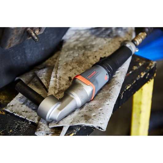 A PROTO J150WA pneumatic torque wrench lies on a dirty cloth on a workbench in a workshop setting. The tool features a grey and black handle with orange accents and is connected to an air hose.