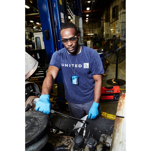 PROTO J5450 red tool bag on floor in workshop background near technician in blue gloves using a ratchet wrench.