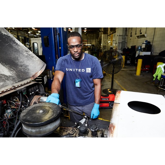 PROTO J5450 ratchet wrench used by a mechanic with blue gloves working on an engine in a workshop, tool bag in background.