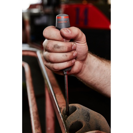 Photo of a hand using a PROTO JD1402R screwdriver with a gray and red handle on a dusty metal surface.